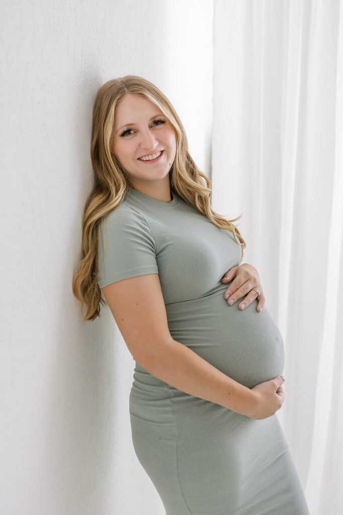 Relaxed maternity portrait of mom in a casual green dress leaning gracefully against a white studio wall.