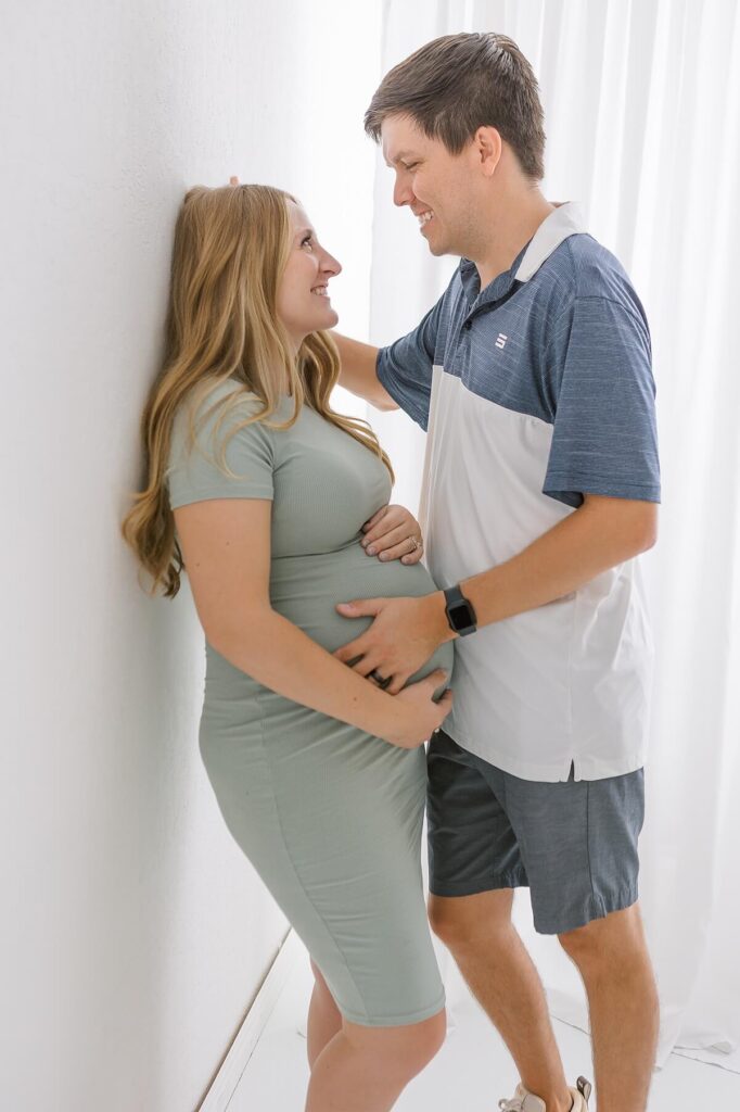 Relaxed maternity portrait of mom in a casual green dress leaning gracefully against a white studio wall.