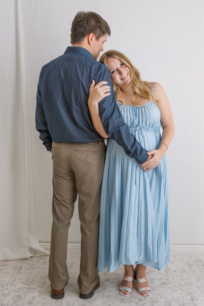 Maternity couple portrait with mom in a blue gown leaning on dad’s arm, both smiling in a white studio setting.