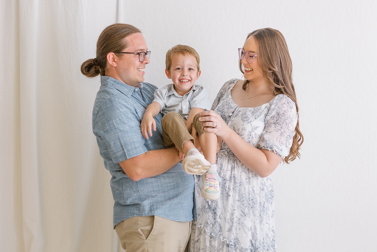 Family of three in studio maternity session in mesa arizona with mom and dad looking at their toddler son and they are all smiling.