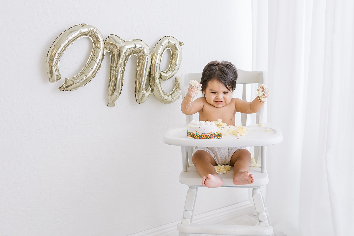 little boy with lots of dark hair sitting in white high chair smashing a cake for his first birthday.