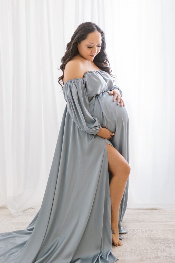 beautiful maternity photo of a pregnant woman standing in white studio in phoenix with glowing light behind her in a flowy blue dress.